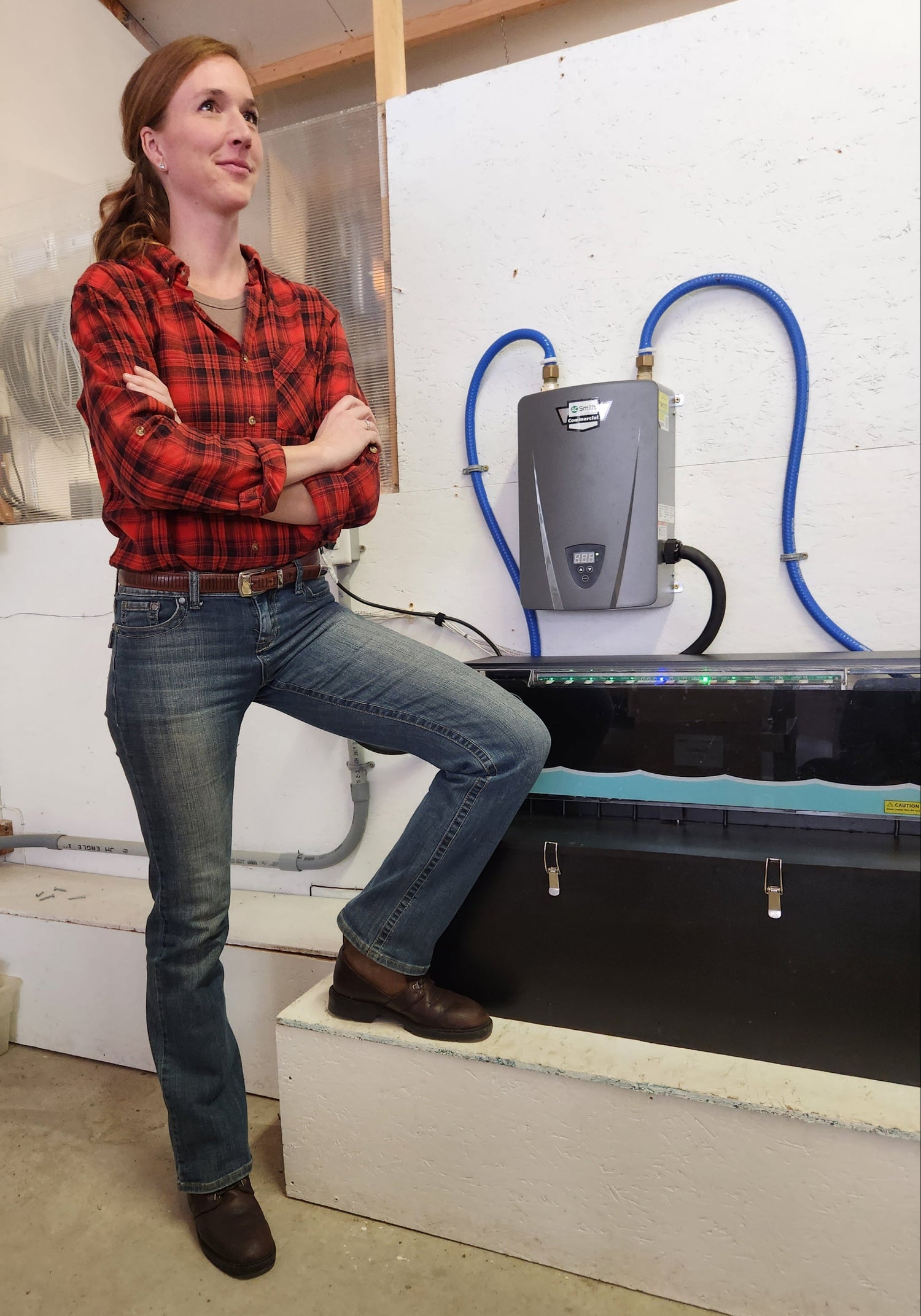 Steph is standing in a proud pose next to the HUG Hydronics system she just installed.  Her hands are folded and her right leg is up near the tank in floor heating system. The tank is suitcase sized and black and has an electric tankless water heater just above it connected by blue hoses. The lights are on top of the tank glowing blue and green indicating it is working well.