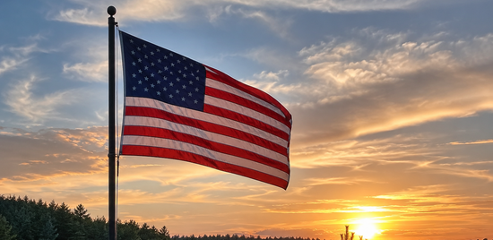 the united states flag flying over a pine treed lake shore line during a sunset